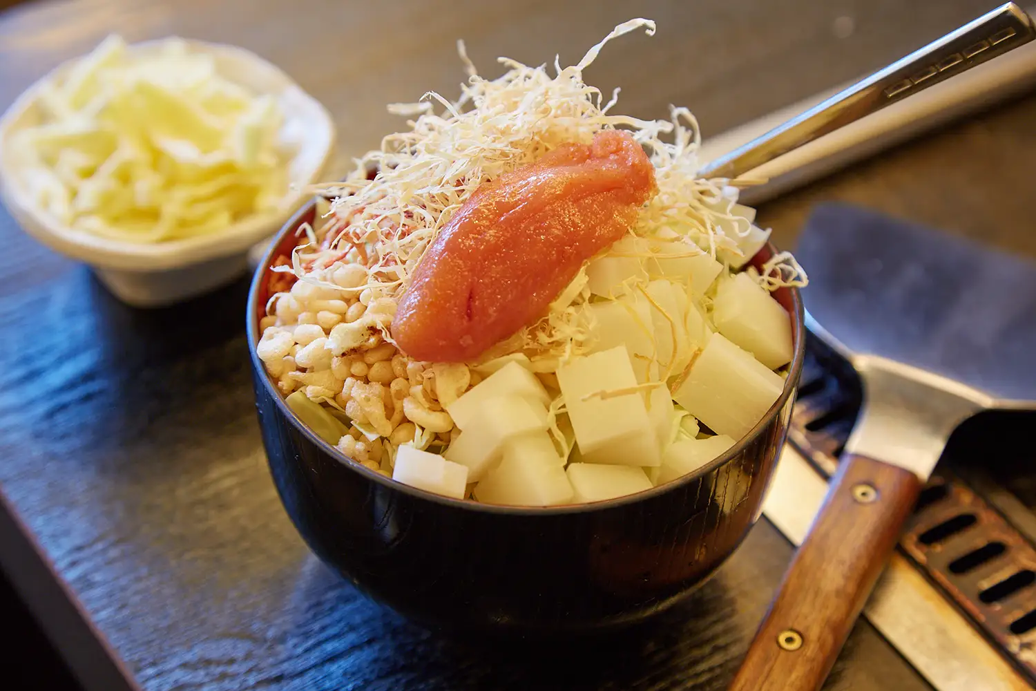 鉄板の横に置かれた、黒いどんぶりに入ったもんじゃ焼きの生地。器に餅、天かす、明太などが大盛りに盛られている。