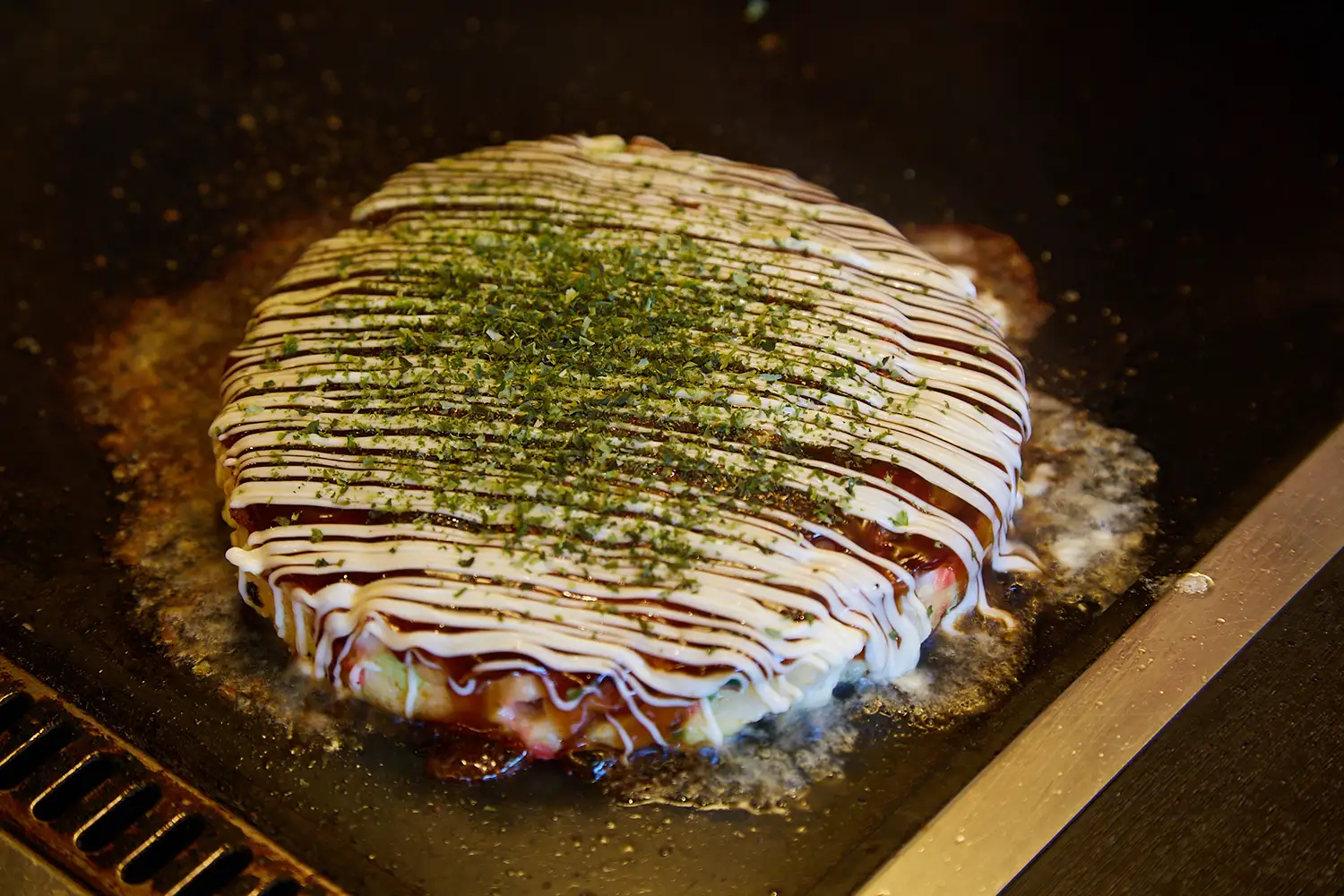 鉄板で焼かれたお好み焼き。ソースとマヨネーズ、青のりがかかっている。