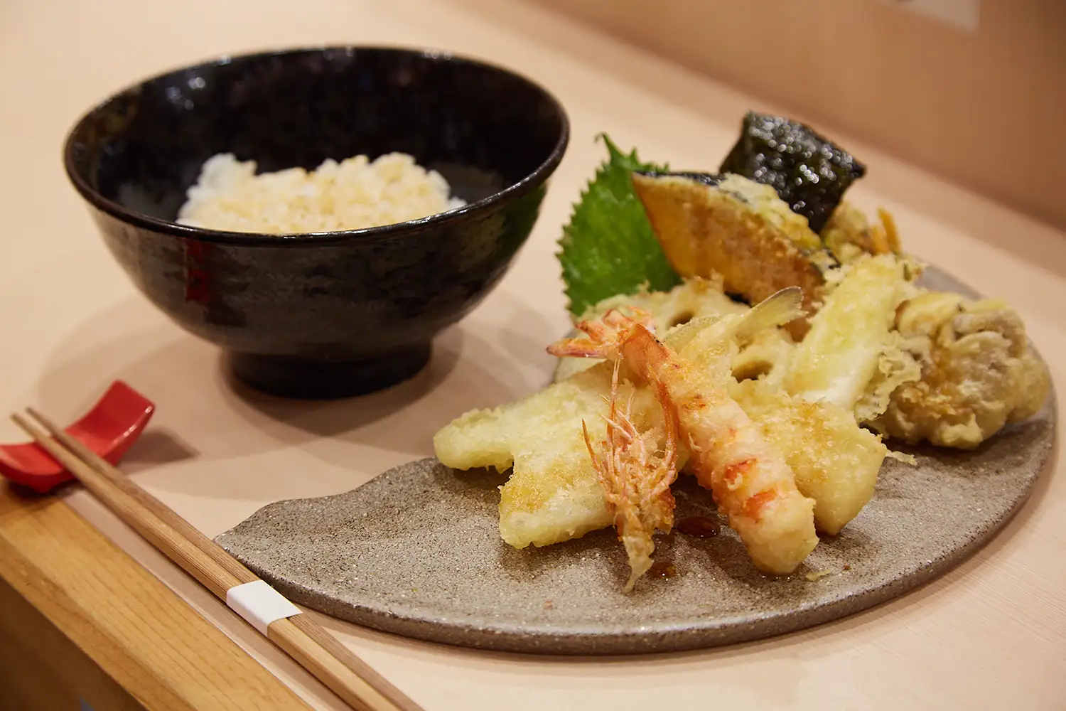黒い茶碗と、平皿に盛られた天ぷら。海老や白身魚、レンコンなどが上品に盛り付けられている。