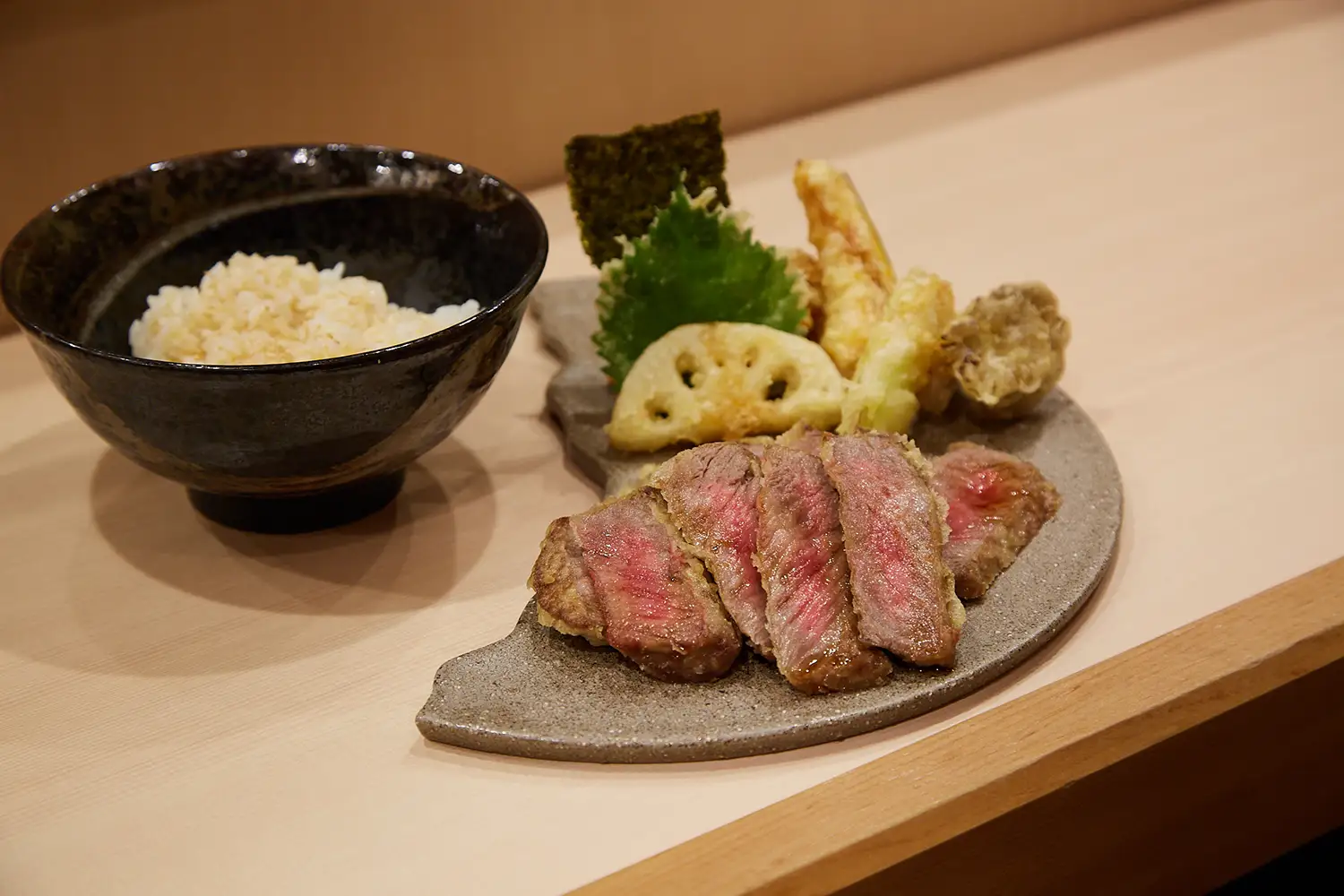 和牛ぜいたく天丼の具材。和牛の薄切り肉と、天ぷら（レンコン、海苔、大葉、野菜など）が皿に盛り付けられ、隣に米飯が入った黒い茶碗が置かれている。