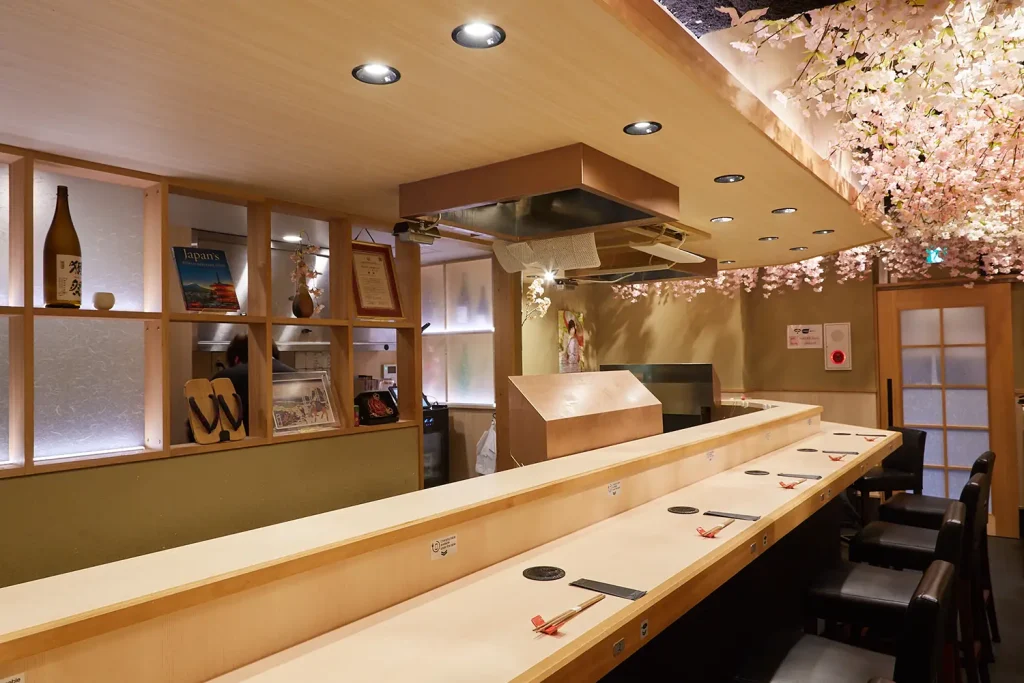 Tempura Asakusa SAKURAの店内にあるカウンター席。天井には桜の造花が広がり、左手の棚には酒瓶や飾り物が置かれている。